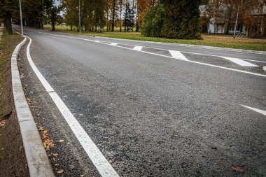 Sessiz, boş bir asfalt yol, beyaz bir ok ve şerit işaretleri. Sahne, arka plandaki yaprakları ve ağaçları gösteriyor. Yakındaki bina kısmen görülebiliyor..