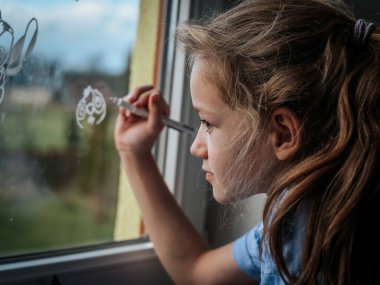 Genç bir kız beyaz kalemle pencereye resim çiziyor. Çizgi film karakteri yaratmaya odaklanıyor. Doğal gün ışığı onun yaratıcılığını ve konsantrasyonunu ön plana çıkarıyor..