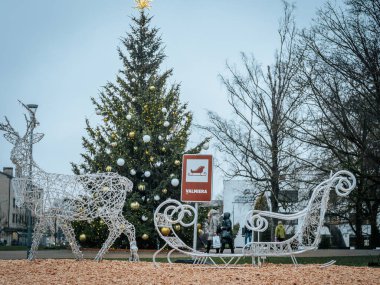 A festive open-air exhibition with a wire reindeer and sleigh sculpture on wood chips, a decorated Christmas tree with gold and white ornaments, and a sign with the inscription 