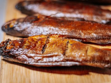 Zengin kahverengi derisi ahşap bir yüzey üzerinde duran tütsülenmiş balığın detaylı bir yakın çekimi. Balığın dokusu ve ayrıntıları açıkça görülüyor, pişirme sürecini vurguluyor..