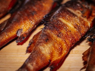 Zengin kahverengi derisi ahşap bir yüzey üzerinde duran tütsülenmiş balığın detaylı bir yakın çekimi. Balığın dokusu ve ayrıntıları açıkça görülüyor, pişirme sürecini vurguluyor..