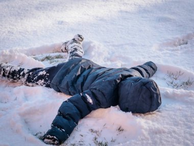 Kışlık ceketli küçük bir çocuk, kolları ve bacakları açık, karda sırt üstü uzanmış, parlak kış güneşi altında bir kar meleği yaratıyor..