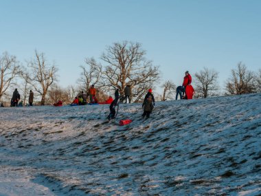 Bir grup insan, açık mavi bir dağın altında yapraksız ağaçlarla çevrili karlı bir dağda kızak ve açık hava aktivitelerini seviyor. 