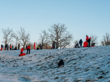 Bir grup insan, açık mavi bir dağın altında yapraksız ağaçlarla çevrili karlı bir dağda kızak ve açık hava aktivitelerini seviyor. 