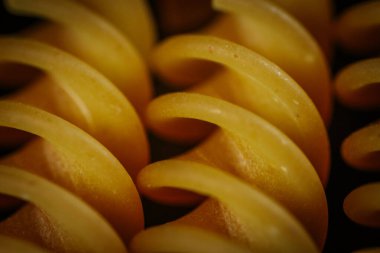 Aşırı yakın plan sarı rotini makarna sarmalı, düz dokularını ve kıvrımlı şekillerini koyu arkaplana karşı detaylı bir kompozisyonda gösteriyor..