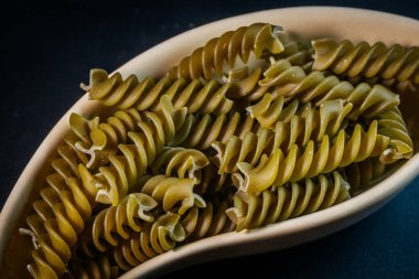 Koyu arkaplana yerleştirilmiş bej bir seramik kasede pişmemiş yeşil rotini makarna sarmalı. Resim dokuyu ve ayrıntıyı vurgular.