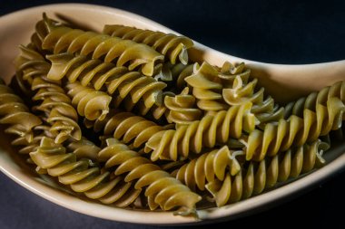 Koyu arkaplana yerleştirilmiş bej bir seramik kasede pişmemiş yeşil rotini makarna sarmalı. Resim dokuyu ve ayrıntıyı vurgular.