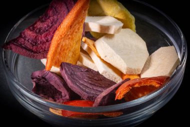 Çeşitli kurutulmuş sebze ve meyve dilimlerinin yakın plan çekimi. Bir cam kasede mor tatlı patates, havuç ve diğer parlak parçalarla birlikte..