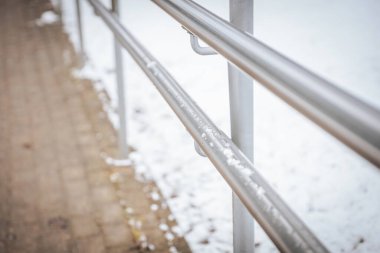 Üzerinde hafif kar tozu olan metal bir tırabzanın yakın çekimi asfalt kaldırım boyunca uzanıyor. Bulanık karlı arka plan derinlik ve kış atmosferi ekler..