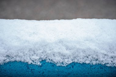 Yeni yağmış karla kaplı mavi metal korkulukların yakın çekimi. Karların yumuşak dokusu korkuluğun sert yüzeyiyle çelişiyor. Bulanık arkaplan derinlik ekler.