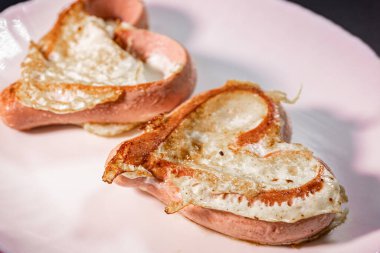 Beyaz bir tabakta kızarmış yumurtayla doldurulmuş kalp şeklinde sosisler. Tabak, yaratıcı ve lezzetli bir kahvaltı sunumu için altın sarısı kıtır kıtır bir dokuya sahip..