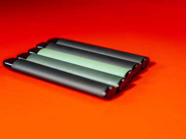 Parlak turuncu arka planda kesişen iki yeşil e-sigara kaleminin yakın çekimi. Parlak tasarım ve kalın renkler çarpıcı bir görsel karşıtlık yaratır.