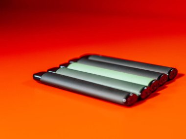 Parlak turuncu arka planda kesişen iki yeşil e-sigara kaleminin yakın çekimi. Parlak tasarım ve kalın renkler çarpıcı bir görsel karşıtlık yaratır.