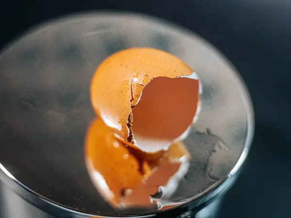 Yansıtıcı metal bir yüzey üzerinde duran kırık kahverengi yumurta kabuğunun yakın çekimi. Kırık kabuk, koyu arkaplanla çelişen bir ayna görüntüsü yaratır..