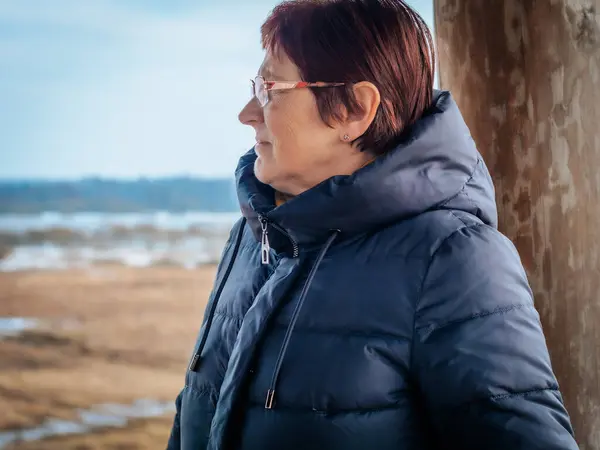 Orta yaşlı, gözlüklü ve koyu mavi ceketli bir kadın tahta bir direğin yanında duruyor ve uzaklığa bakıyor. Bulanık orman arka planı doğal ve barışçıl bir atmosfer yaratır..