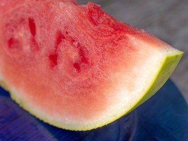 Taze karpuz diliminin makro görüntüsü, sulu kırmızı etini, yeşil kabuğunu ve doğal dokusunu gösteriyor. Canlı meyve mavi bir tabakta oturur ve tazeleyici cazibesini vurgular..