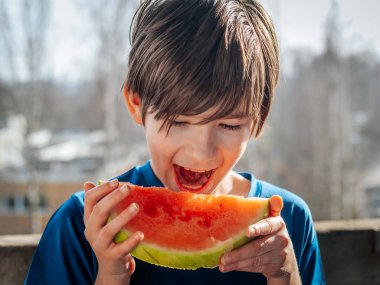 Islak saçlı genç bir çocuk güneşli bir günün tadını çıkarırken taze bir karpuzu ısırıyor. Ağaçlar ve binalar bulanık arka planda görülebilir, görüntüye derinlik verir.