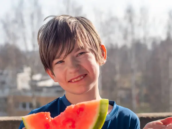Islak saçlı genç bir çocuk güneşli bir günün tadını çıkarırken taze bir karpuzu ısırıyor. Ağaçlar ve binalar bulanık arka planda görülebilir, görüntüye derinlik verir.