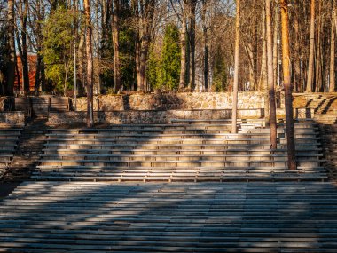 Uzun ağaçlarla çevrili açık hava amfi tiyatrosunda sıra sıra yıpranmış ahşap banklar yer alıyor. Sıcak güneş ışığı oturma alanına uzun gölgeler düşürüyor ve huzurlu bir atmosfer yaratıyor..