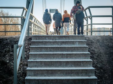 Bir grup insan beton bir merdiven boşluğundan görünen yaya köprüsünden geçiyor. Metal parmaklıklar ve kentsel zemin doğal çevreyle tezat oluşturuyor. 03.09.2025 Valmiera, Letonya