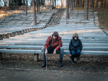 İki çocuk açık hava amfi tiyatrosunda aşınmış ahşap banklarda oturuyor. Biri kırmızı bir ekose ceket giyer ve bir sopayla oynar, diğeri ise kapüşonlu bir ceket giyer, aşağı bakar. Ortalık sessiz ve düzensiz.