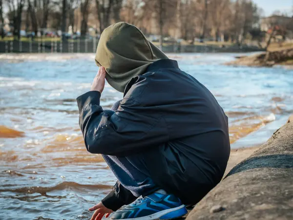 Kapüşonlu ceketli genç bir çocuk nehrin kenarında oturur ve sakince kameraya bakar. Doğal ışık yüzüne vurgu yaparken, arka planda akan su derinlik katıyor..
