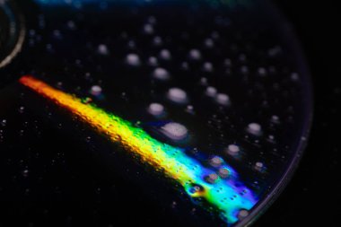 İçinde su damlacıkları olan bir CD 'nin yakın çekimi yumuşak ışıktaki yanardöner yansımaları ortaya çıkarıyor. Koyu arkaplan parlak gökkuşağı renkleri ve karmaşık dokularla çelişiyor..