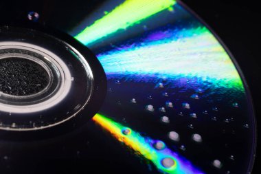 İçinde su damlacıkları olan bir CD 'nin yakın çekimi yumuşak ışıktaki yanardöner yansımaları ortaya çıkarıyor. Koyu arkaplan parlak gökkuşağı renkleri ve karmaşık dokularla çelişiyor..