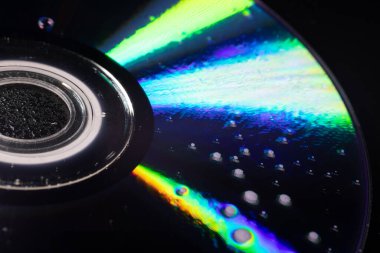 İçinde su damlacıkları olan bir CD 'nin yakın çekimi yumuşak ışıktaki yanardöner yansımaları ortaya çıkarıyor. Koyu arkaplan parlak gökkuşağı renkleri ve karmaşık dokularla çelişiyor..