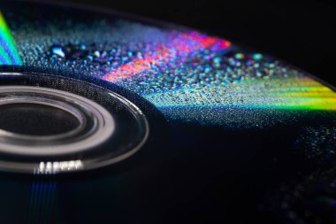 İçinde su damlacıkları olan bir CD 'nin yakın çekimi yumuşak ışıktaki yanardöner yansımaları ortaya çıkarıyor. Koyu arkaplan parlak gökkuşağı renkleri ve karmaşık dokularla çelişiyor..
