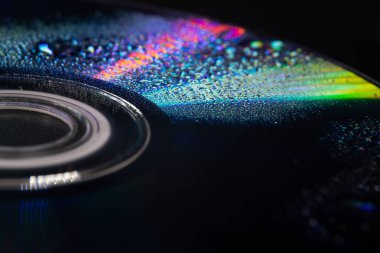 İçinde su damlacıkları olan bir CD 'nin yakın çekimi yumuşak ışıktaki yanardöner yansımaları ortaya çıkarıyor. Koyu arkaplan parlak gökkuşağı renkleri ve karmaşık dokularla çelişiyor..