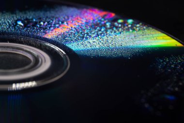 İçinde su damlacıkları olan bir CD 'nin yakın çekimi yumuşak ışıktaki yanardöner yansımaları ortaya çıkarıyor. Koyu arkaplan parlak gökkuşağı renkleri ve karmaşık dokularla çelişiyor..