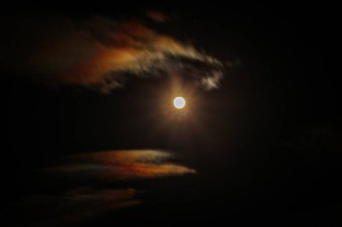 Dolunay gece gökyüzünde parlıyor, turuncu ve mavi renkli yanardöner bulutlarla çevrili, mistik ve atmosferik bir manzara yaratıyor..