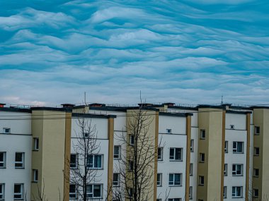 Bej renkli bir apartman dairesi dramatik bir gökyüzünün altında duruyor. Önünde ağaçlar ve elektrik hatları olan mavi bulutlarla dolu..