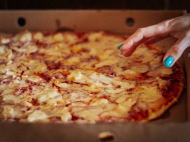 Karton kutuda peynirli çivi ve jambonlu pizza için uzanan mavi boyalı bir el. Pizza dilimlenmemiş, eritilmiş malzemeler ve ince bir kabukla..
