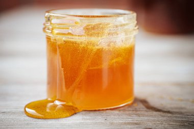 Kenarlarından taşralı ahşap bir yüzeye dökülen altın bal dolu cam bir kavanozun yakın çekimi. Bal, kehribar renginde parıldıyor..