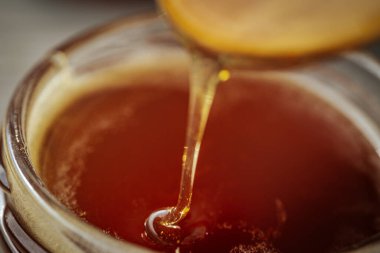 Bir kaşıktan cam bir kavanoza damlayan altın bal, zengin, pürüzsüz dokusunu ve doğal rengini gözler önüne seriyor. Sıcak tonlar kompozisyona hakim..