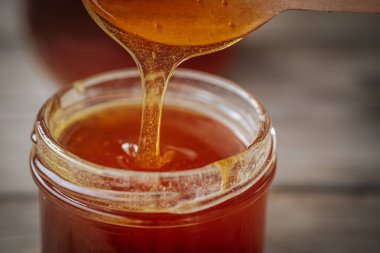Bir kaşıktan cam bir kavanoza damlayan altın bal, zengin, pürüzsüz dokusunu ve doğal rengini gözler önüne seriyor. Sıcak tonlar kompozisyona hakim..