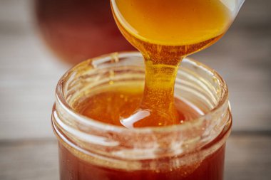 Bir kaşıktan cam bir kavanoza damlayan altın bal, zengin, pürüzsüz dokusunu ve doğal rengini gözler önüne seriyor. Sıcak tonlar kompozisyona hakim..