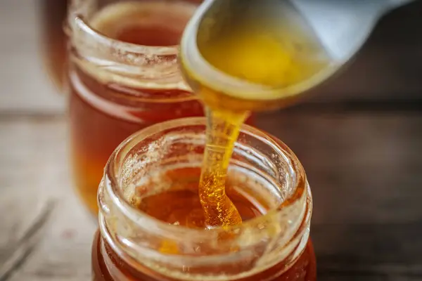 Bir kaşıktan cam bir kavanoza damlayan altın bal, zengin, pürüzsüz dokusunu ve doğal rengini gözler önüne seriyor. Sıcak tonlar kompozisyona hakim..