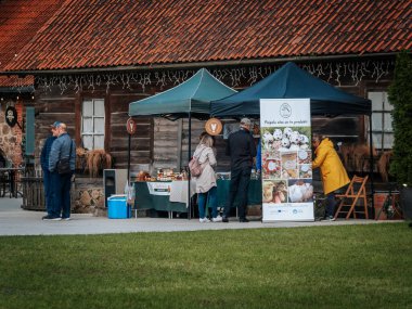 İki kadın kırsal bir pazarda kavanoz ve çiftlik ürünleri sergileyerek konuşuyor. Arkalarında mavi takım elbiseli bir satıcı var. Posterde bıldırcın ve kümes hayvanı ürünleri var..