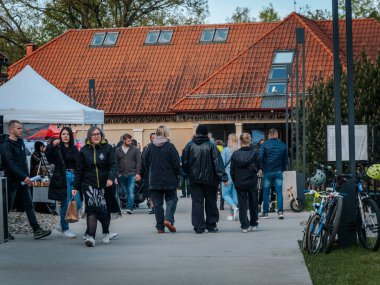 Kalabalık, kırmızı kiremitli çatısı olan bir binanın yakınındaki açık hava pazarında geziniyor. Kasklı bisikletler yakına park edilmiş ve yol boyunca satıcıların çadırları kurulmuş..