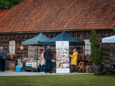 İki kadın kırsal bir pazarda kavanoz ve çiftlik ürünleri sergileyerek konuşuyor. Arkalarında mavi takım elbiseli bir satıcı var. Posterde bıldırcın ve kümes hayvanı ürünleri var..