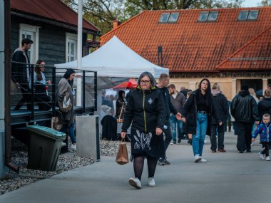 Kalabalık, kırmızı kiremitli çatısı olan bir binanın yakınındaki açık hava pazarında geziniyor. Kasklı bisikletler yakına park edilmiş ve yol boyunca satıcıların çadırları kurulmuş..