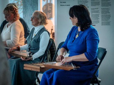 Geleneksel giysiler içinde bir grup kadın, kültürel bir mekanda ahşap ip enstrümanlarıyla gösteri yapıyorlar. Sayfa notaları ve işaretler resmi ya da tarihi bir olayı gösterir.