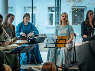 Geleneksel giysiler içinde bir grup kadın, kültürel bir mekanda ahşap ip enstrümanlarıyla gösteri yapıyorlar. Sayfa notaları ve işaretler resmi ya da tarihi bir olayı gösterir.