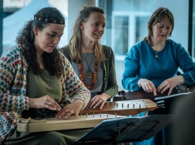 Geleneksel giysiler içinde bir grup kadın, kültürel bir mekanda ahşap ip enstrümanlarıyla gösteri yapıyorlar. Sayfa notaları ve işaretler resmi ya da tarihi bir olayı gösterir.