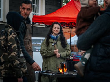 İnsanlar halka açık bir etkinlik için toplanırken, gülümseyen bir kadın küçük bir demir ocağının yanında çekiç tutuyor. Demirhanede kırmızı bir çadır ve bir yangın canlı gösteri sahnesini aydınlatıyor..