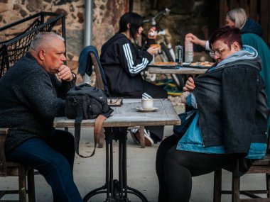 İnsanlar açık bir kafede kırsal ahşap masalarda oturur, bazıları kahve içerken diğerleri pizza yer. Ön plandaki sahne dalgındır..