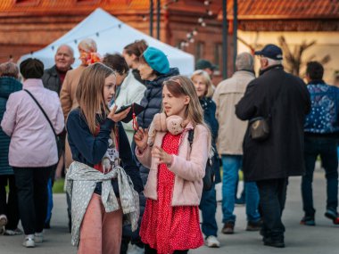 İki genç kız yetişkinler arasında kalabalık bir açık hava etkinliğinde yürüyor. Birinin siyah bir sırt çantası ve sevimli tasarımları var. Arka planda çadırlar ve canlı halk etkileşimi yer alıyor.